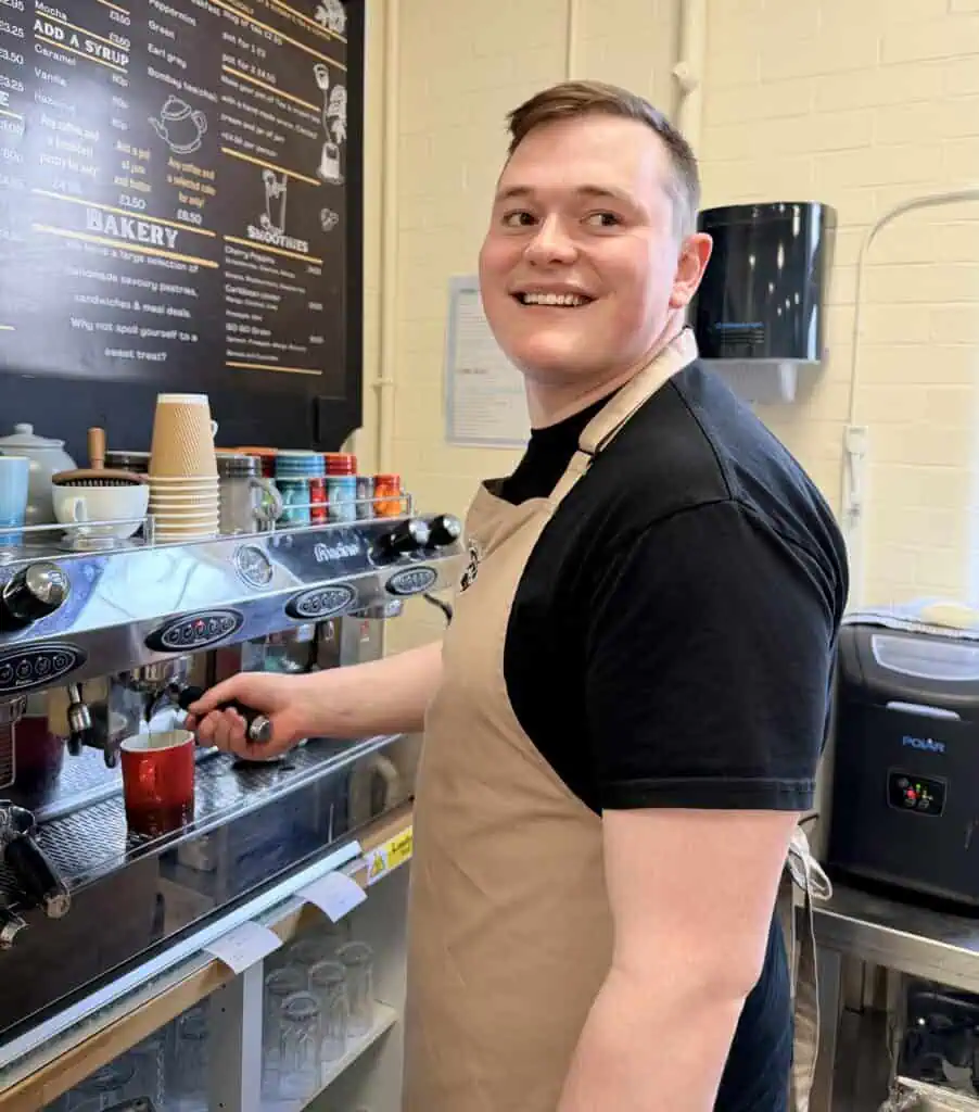 Bright and cheerful atmosphere, the barista wears a beige apron over a black t-shirt, ready to serve fresh coffee.