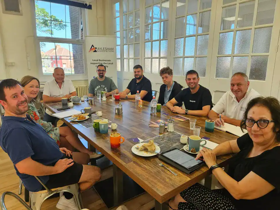 A diverse group of professionals gathered around a wooden table in a bright room with large arched windows, participating in a business meeting for local enterprise support, with coffee, snacks, and laptops.