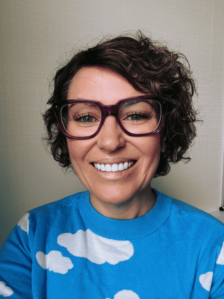 Portrait of Jo Godden smiling, wearing purple glasses and a blue sweater with white cloud patterns.